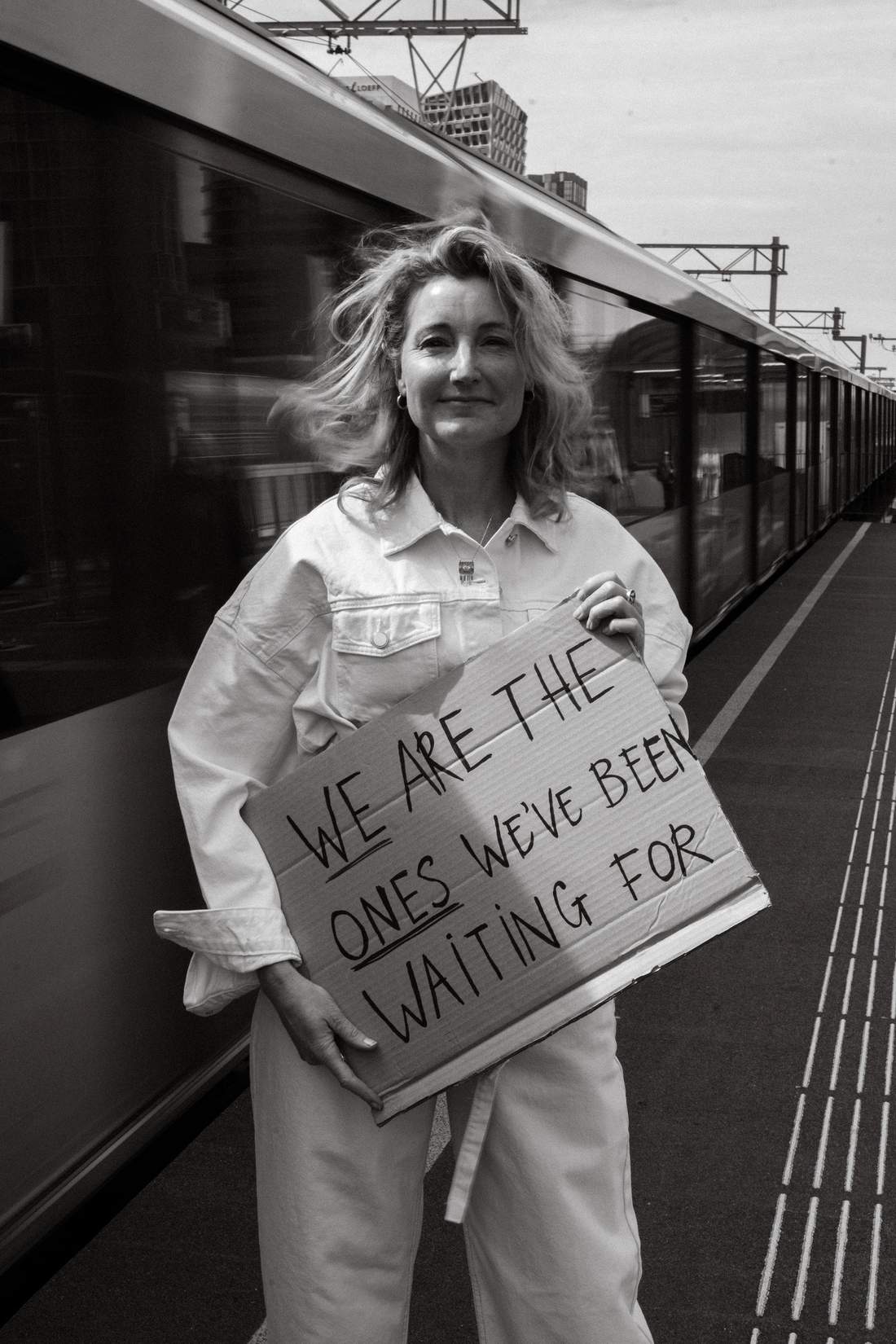 Dian op een perron naast een rijdende trein, met een kartonnen bord: We are the ones we’ve been waiting for