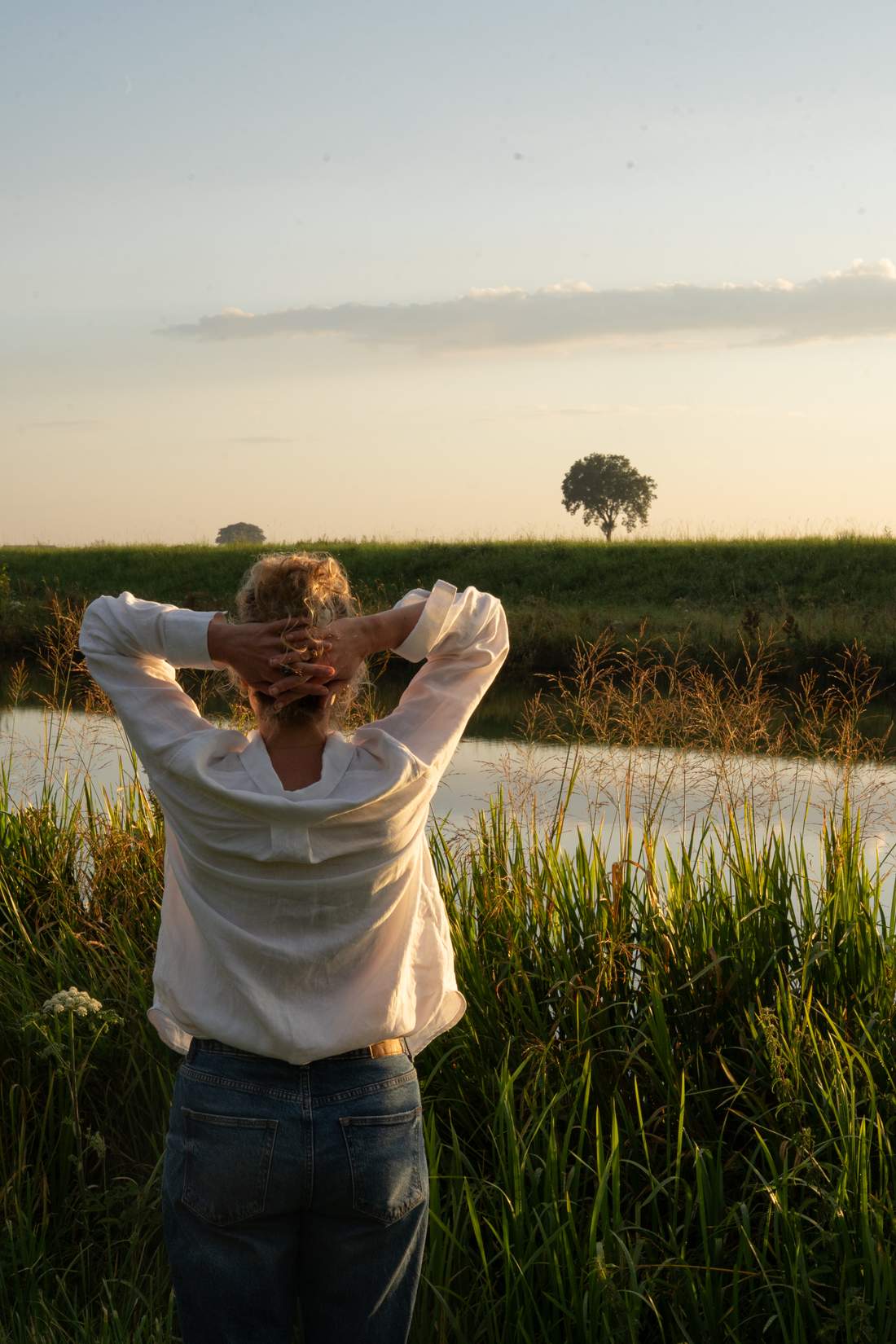 Dian staat met haar rug naar de camera en haar handen achter het hoofd langs het water bij avondlicht